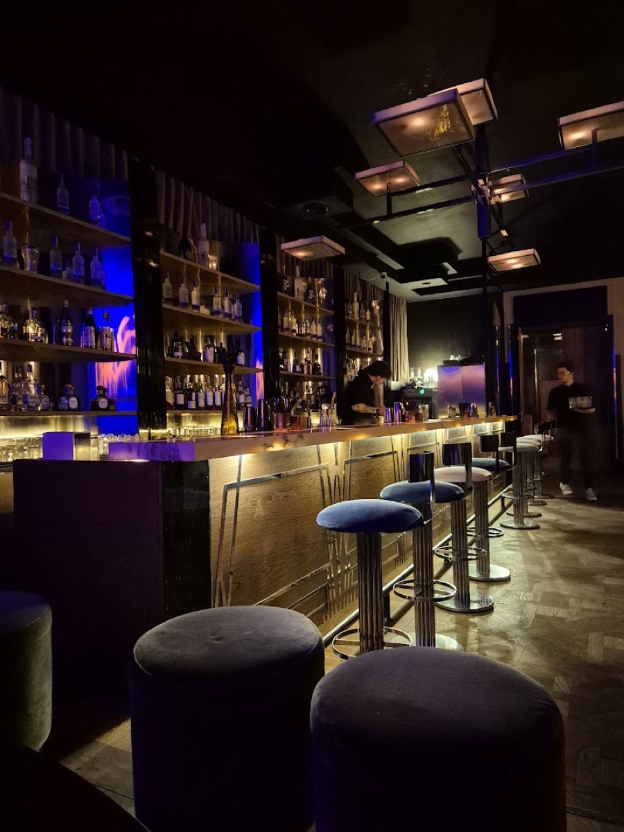 Bar CHARLIE skąpany w błękitnym świetle, z aksamitnymi stołkami i podświetlonymi półkami z butelkami